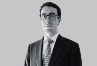 Professional headshot of a man in a dark suit, white shirt, and patterned tie, wearing glasses.